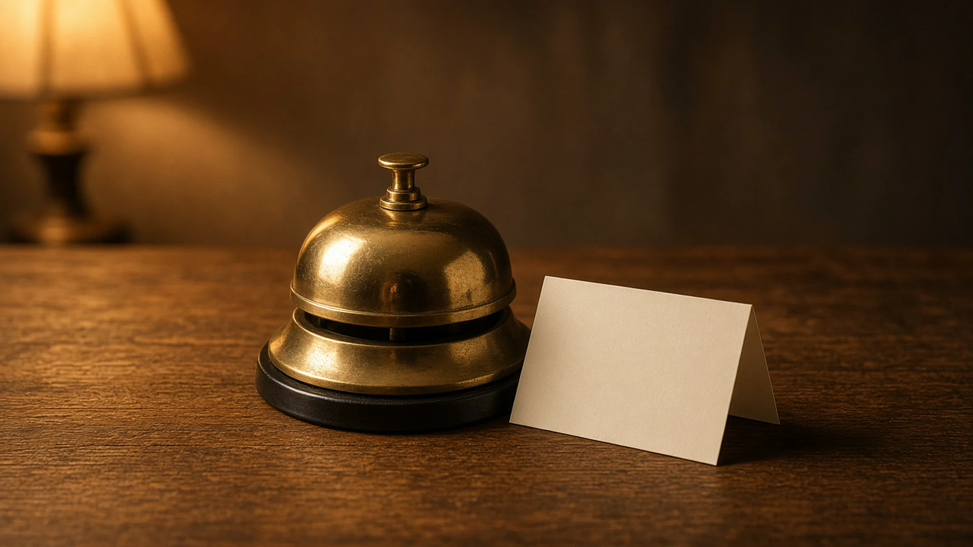 Brass desk bell with a blank folded note beside it on a wooden desk under warm lamp light