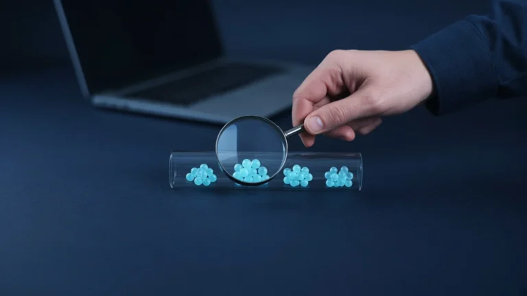 Hand holding a magnifying glass over blue glowing beads flowing through a clear glass pipe on a desk