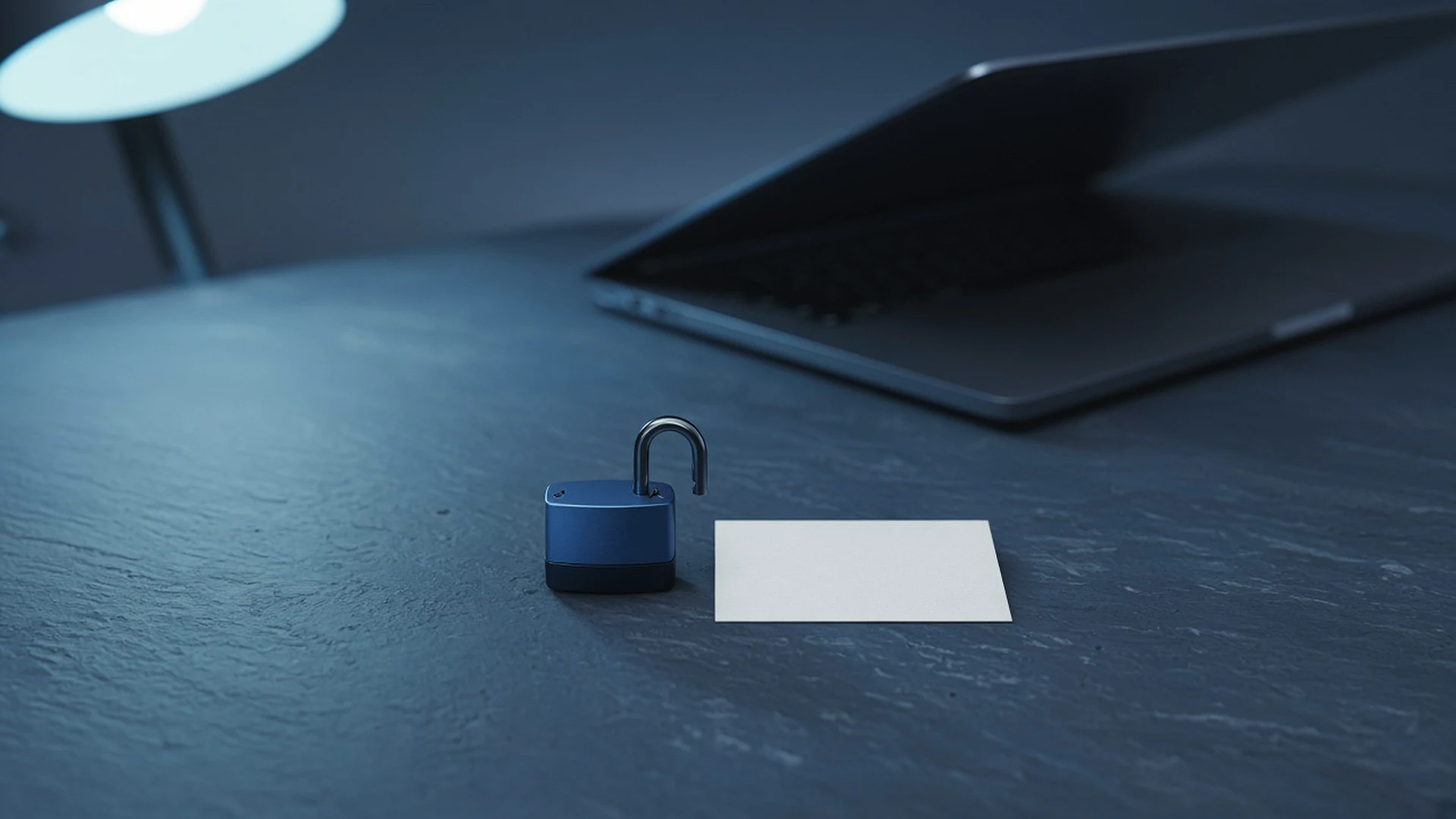 Open steel blue padlock on a desk beside a blank index card and a laptop under cool lamp light